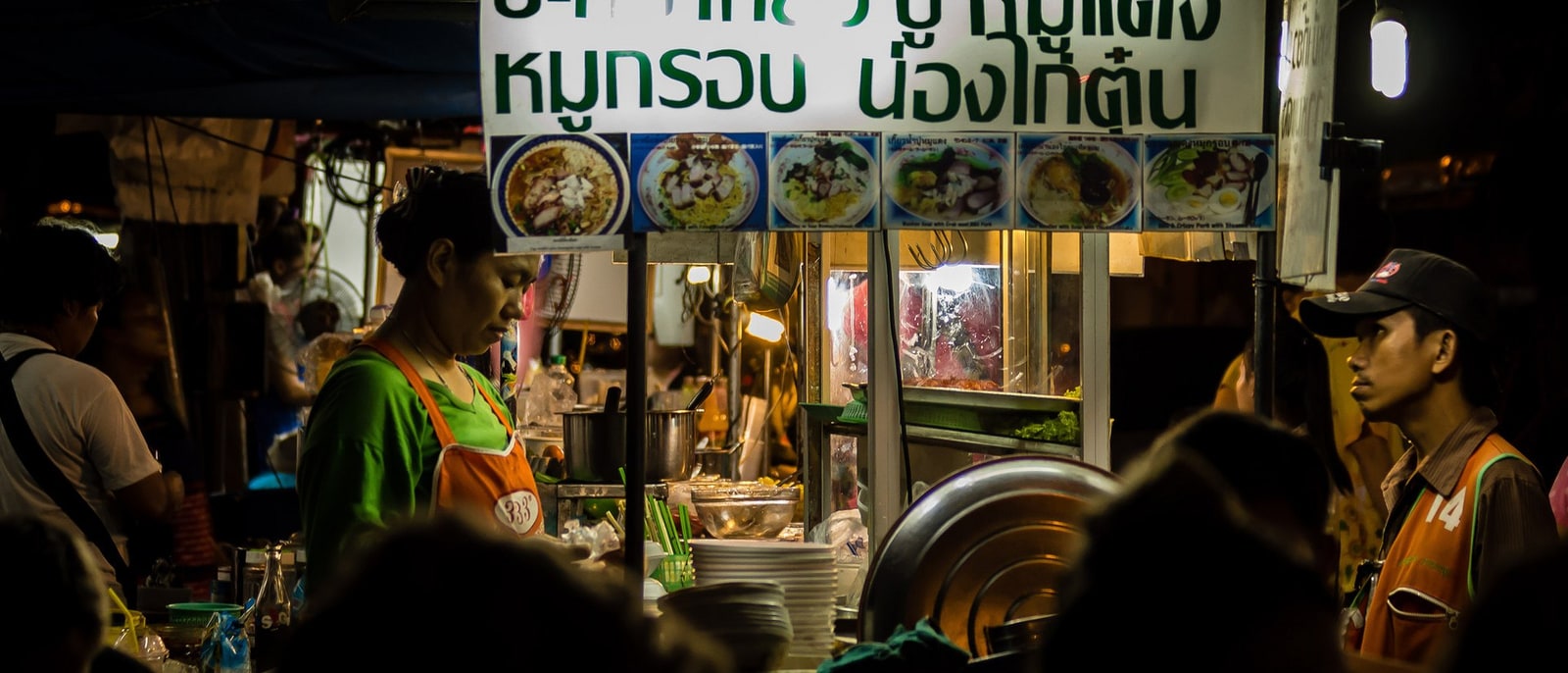 Un puesto de comida callejera de noche en Bangkok, con el cartel iluminado de fotos de los platos y la cocinera al wok