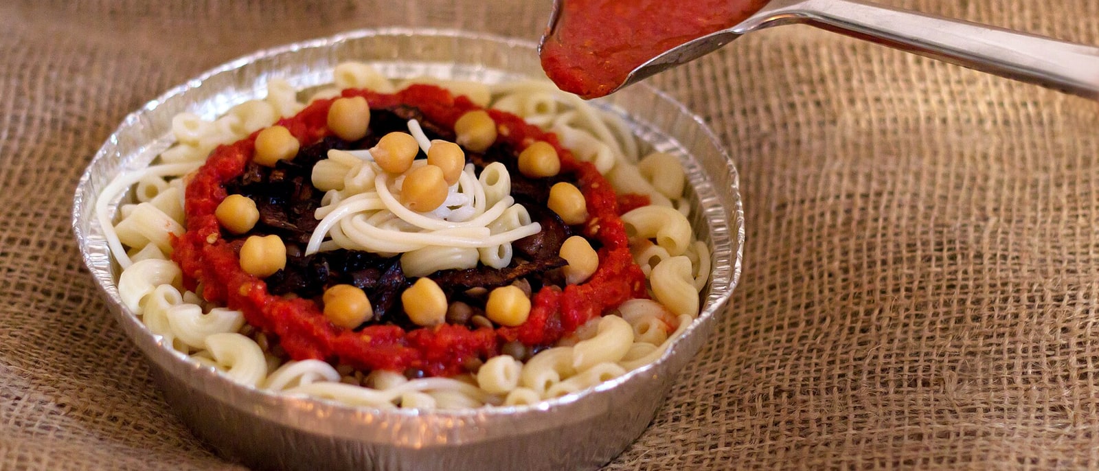 Un koshari servido en su bandeja de aluminio, con la salsa por encima