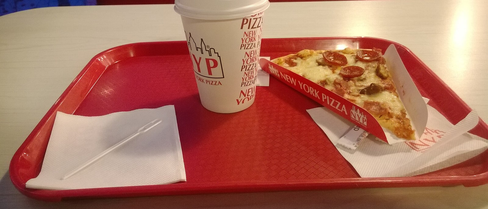 A pizza slice on its paper tray, a cup beside it