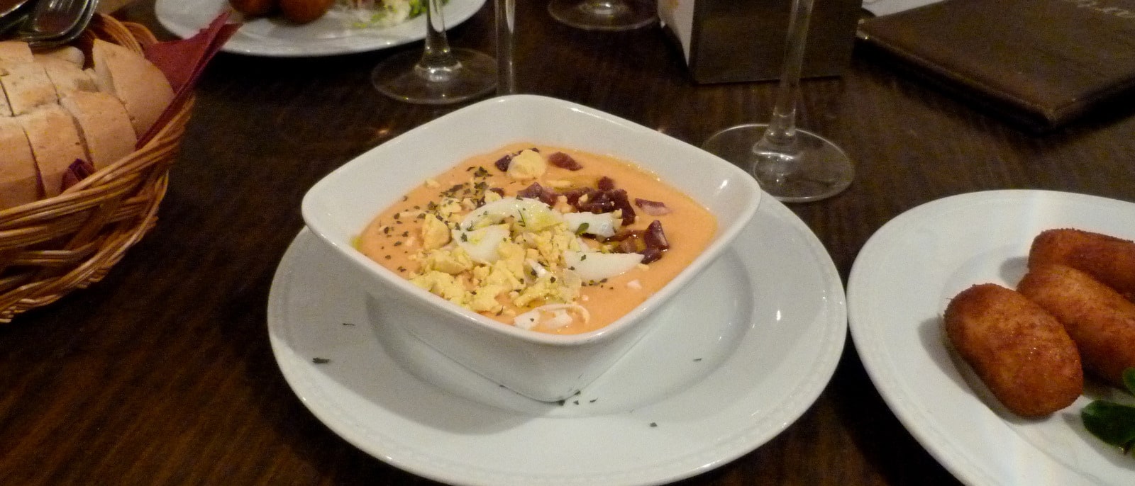 A bowl of salmorejo topped with ham and egg, croquettes and bread beside it