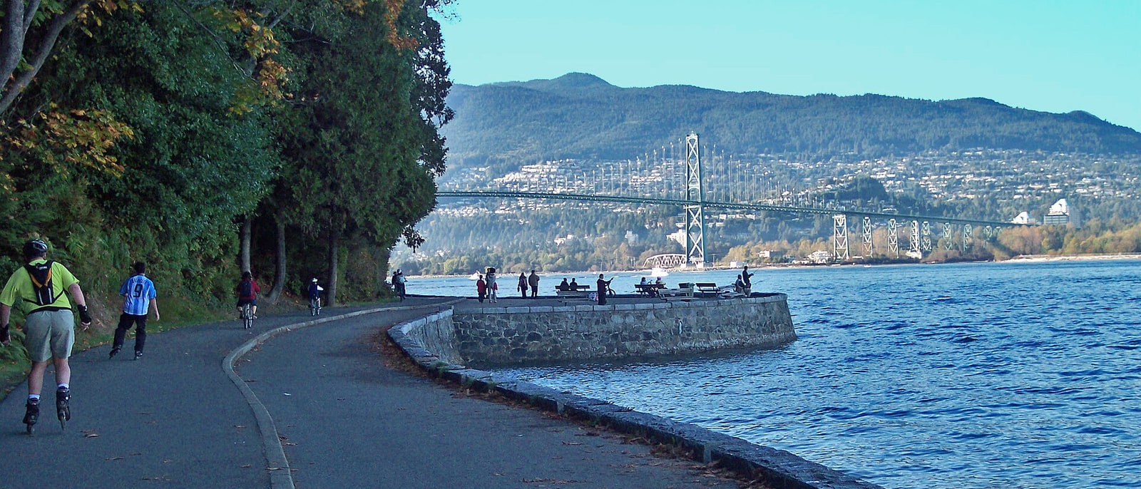 Cyclists and skaters on the Stanley Park seawall, Lions Gate Bridge in the distance